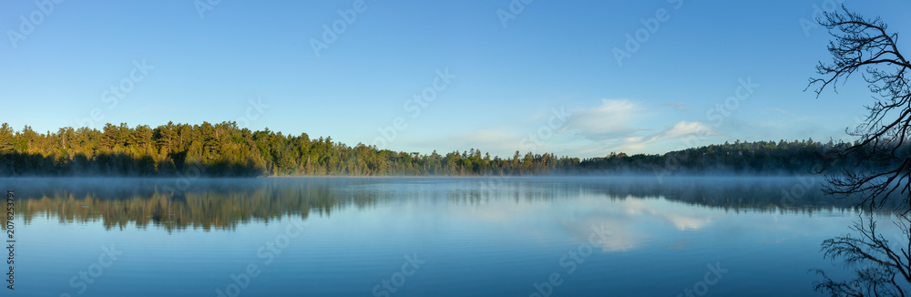 Naklejka premium Panorama of a blue northern Minnesota lake and pines with morning fog under blue sky