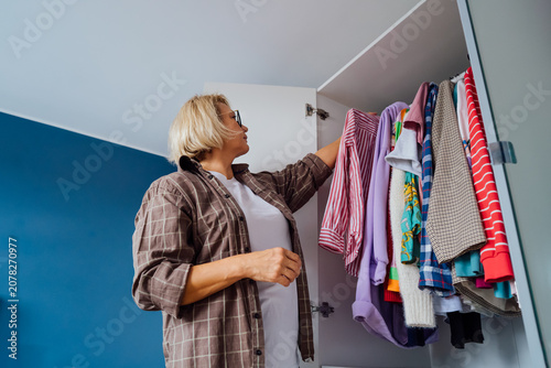 Middle aged woman selecting clothes from wardrobe for donating to Charity shop. Decluttering, Sorting clothes and Cleaning Up. Reuse, second-hand concept. Conscious consumer, sustainable lifestyle.