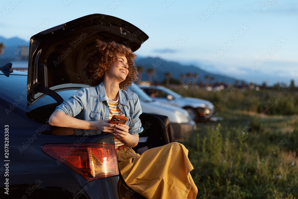 Naklejka premium A smiling woman in a denim jacket sits on a car trunk at dusk, using a smartphone during a relaxed roadside moment, enjoying the calm landscape and casual travel vibe.
