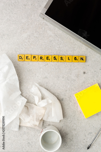 Overhead view of depression awareness concept with letter tiles spelling DEPRESSION, tissues, pill bottle, laptop, sticky note, and pen on a light desk background.