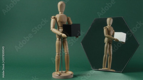 Wooden figure holds black cube, mirror reflects its white counterpart against green backdrop.