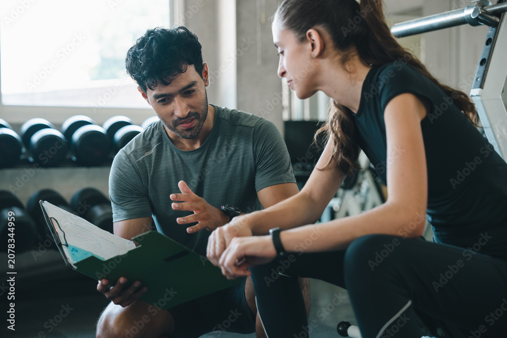 Fototapeta premium Trainer client gym clipboard consultation fitness exercise coaching session woman man discuss workout plan with focused expression near weight rack in modern health club