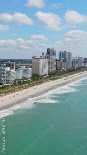 Vertical screen. Scenic coastal view of Myrtle Beach, South Carolina, featuring sandy beach, ocean waves, and high-rise buildings against a bright sky