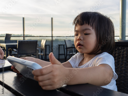 Fotografie A candid portrait of a young girl engrossed in a smartphone at an outdoor cafe terrace, bathed in warm golden-hour light