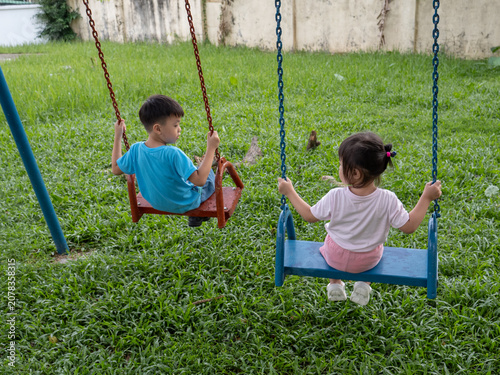 Back view of two young children, a boy and a girl, sitting on red and blue swings in a grassy playground.
