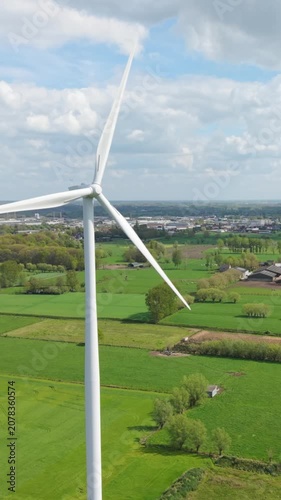 Vertical screen. Aerial perspective of a large wind turbine with rotating blades set in a vibrant green landscape, featuring fields and distant structures under a clear blue sky filled with clouds