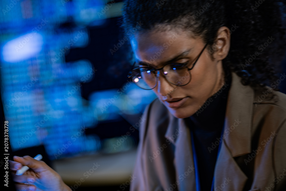 Fototapeta premium A female analyst gazes intently at data off screen, framed against glowing monitors in a dark technology room, in a professional, cutting edge technology setting.