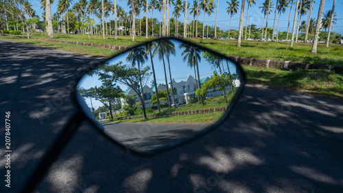 Palm coconut trees line a sunlit tropical road. Mirror reflects a white house and green lawn. Shadows dance across asphalt, hinting at motion. Serene landscape evokes calm, peaceful travel