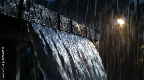 Heavy rain pouring from roof gutter at night with bokeh street light background, cinematic storm atmosphere, and weather concept