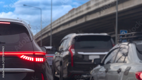 Low angle shot showing the rear of a black car stuck in dense traffic During the day. Turn on brake light. Vehicles are lined up on the highway road beneath a massive concrete overpass structure.