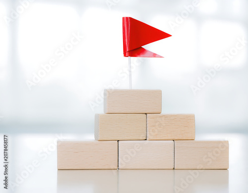 Pyramid of wooden blocks with red flag on top on wooden table with white office shelves background, selective focus and copy space, business development, goal or career success concept AI.