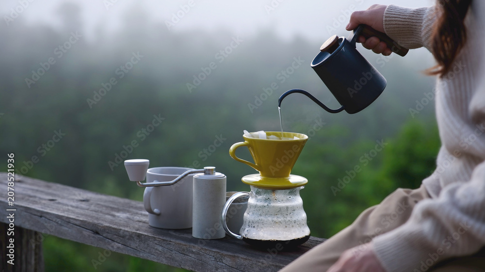 Obraz premium Closeup of a woman making drip coffee on wooden terrace with a mountain view on foggy morning