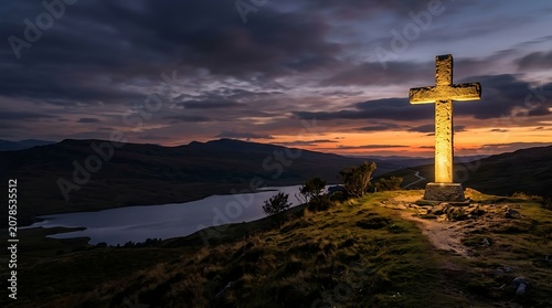 Large illuminated stone cross on a hillside overlooking a scenic lake
