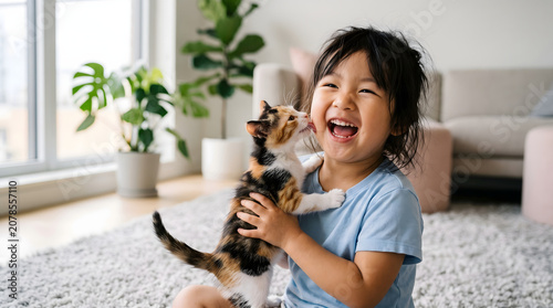 Happy young East Asian girl laughing while holding a cute calico kitten that licks her cheek in a bright living room