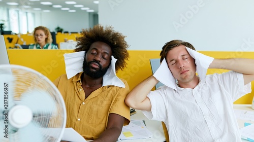 Overheated office workers sitting at desks with an electric fan, showing signs of heat exhaustion and low productivity during a summer heatwave