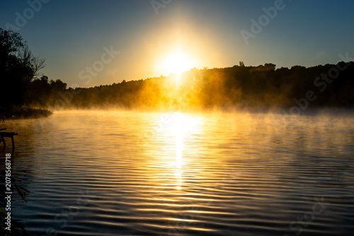 Konstfotografi Early sun rises beyond the wooded hill and reflects in widening ripples while va