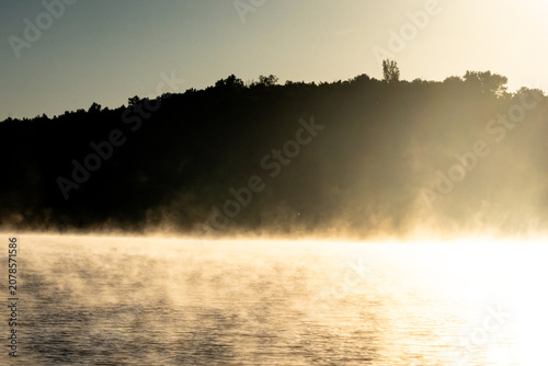 Obraz na płótnie Sunlit mist stretches across the lake below a shadowed hill while the sky remains pale and clean