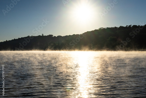 Bright sunrise over the lake sends a shimmering path of light across drifting steam fog. The wooded shore rests in shadow and the water surface rolls with calm ripples and morning hush.