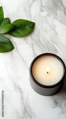 Lit candle in matte black container beside green leaves on white marble surface top view