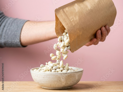 Pouring white beans from a brown paper bag into a ceramic bowl, healthy plant-based cooking ingredients, raw organic navy beans on pink background
