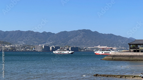 Fototapeta 快晴の青空の下、宮島と対岸を結び瀬戸内海を運航する宮島フェリーの風景、安芸の宮島への交通アクセスと美しい海の景色、広島県観光。