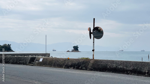 Photos 高松市の海岸沿いの道路とカーブミラー、防波堤の向こうに広がる穏やかな瀬戸内海と行き交う貨物船のノスタルジックな風景。