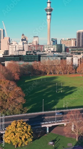AUCKLAND, NEW ZEALAND, 27 June 2026 – An aerial view shows the Sky Tower and city skyline overlooking a rugby field with players practicing and cars driving on a highway below.