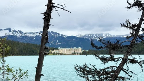 The camera shows a lake with blue water and a historical hotel in the background. Mountains surround the area with some snow on the peaks. Trees line the shore.