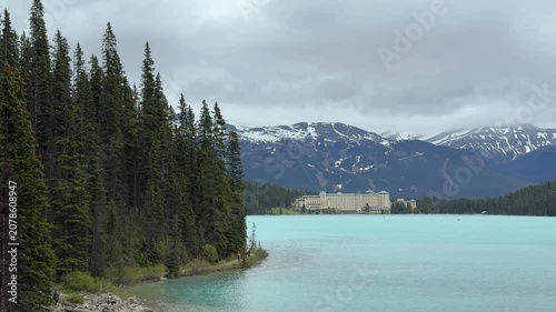The camera shows a lake with blue water and a historical hotel in the background. Mountains surround the area with some snow on the peaks. Trees line the shore.