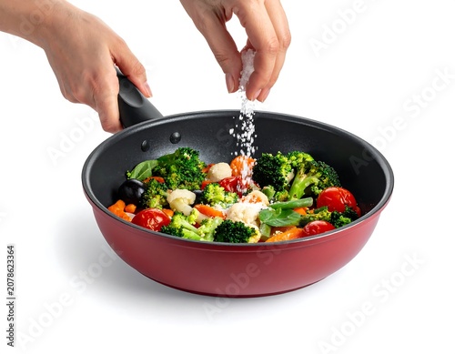 A person seasoning a vibrant mix of vegetables in a red frying pan