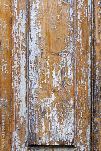 An old wooden door with rectangular carvings is coated with a protective varnish. Peeling white paint is visible in places. The texture and grain of the wood are visible.