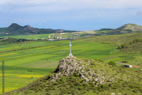 A view of the valley and green meadows, plowed fields. A tall cross on a cliff. Mountains and clouds in the background. Elia, Kakheti, Georgia