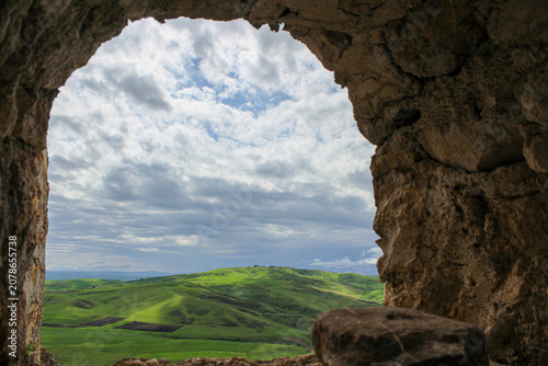 A view of the valley and green meadows through the arch of the monastery wall. Mountains and clouds are in the background. Elia, Kakheti, Georgia