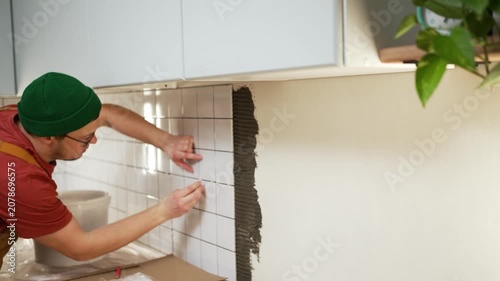 Concentrated male builder in overalls applying small white ceramic tiles on a kitchen wall, using plastic spacers for even joints during a home improvement or renovation project
