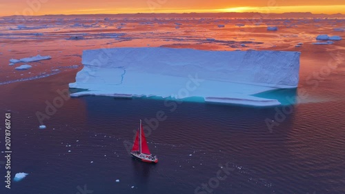 Red sailboat sails near huge iceberg at sunset
