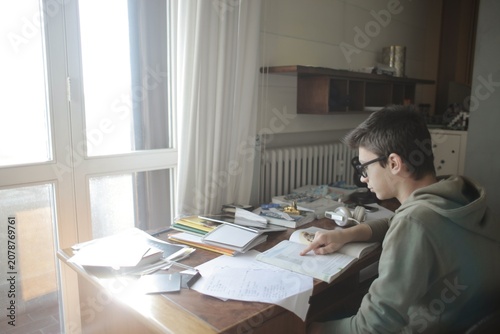 Teenage boy with glasses studying at messy desk by window