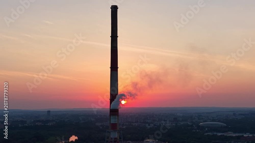 Industrial smokestack emits steam against a vibrant sunset backdrop, showcasing a gradual transition in light and color over a cityscape with trees and buildings below