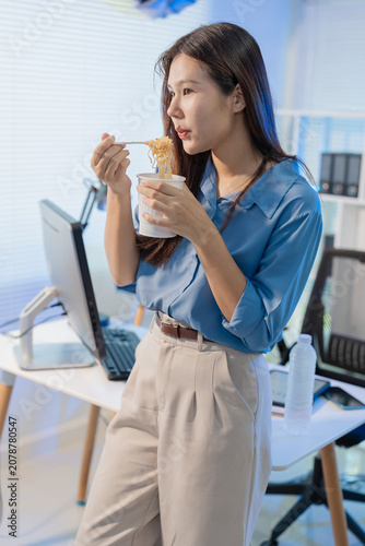 Asian businesswoman eating instant cup noodles while working at her office desk at night.
