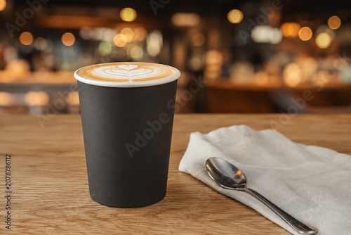 Blank Black Disposable Paper Coffee Cup Mockup with Latte Art on Wooden Table, Featuring Metal Spoon and Napkin in a Warm Blurred Cafe Interior for Branding Presentation.