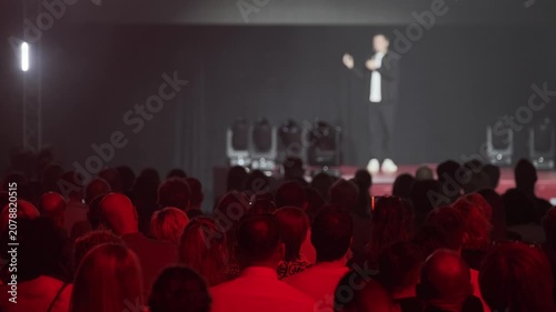 Audience watching speaker on stage at conference, red stage lighting creates moody atmosphere, blurred presenter and attentive crowd.