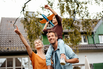 Woman pointing by man carrying playful son on shoulders holding toy airplane in backyard