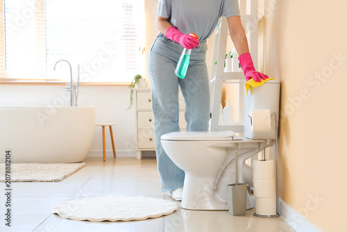 Young woman with rag and spray cleaning toilet in bathroom