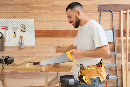 Happy male construction worker with professional tools and handsaw sawing woo...