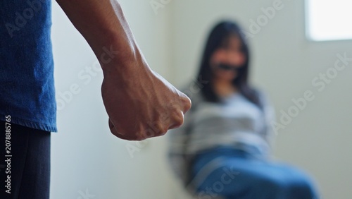 Fotografie Criminal Clenches His Fist In The Foreground While A Scared Young Female Hostage