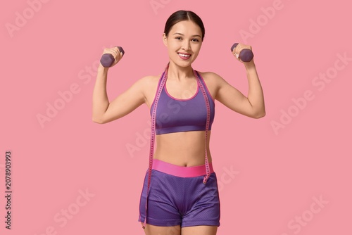 Young woman in sportswear, with measuring tape and dumbbells on pink backgrou...