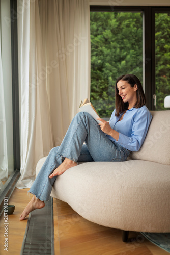 Relaxed woman reading a book while sitting on a comfortable sofa at home