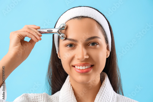 Beautiful young woman massaging her face with tool on blue background, closeup