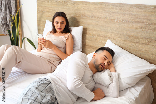 Displeased young woman with mobile phone and her sleeping husband in bedroom