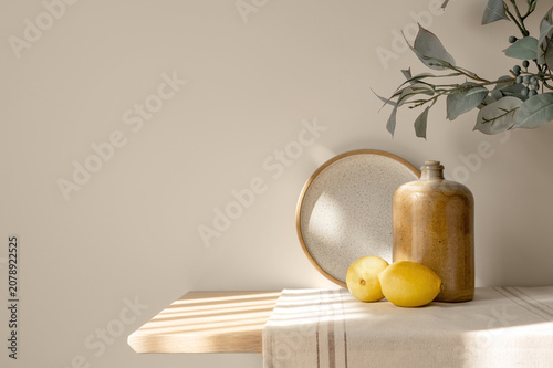 Ceramic dishes, lemons on wooden table with beige linen runner, floral branch, empty warm beige wall background copy space, natural sunlight shadows. Cozy cottage Mediterranean kitchen interior