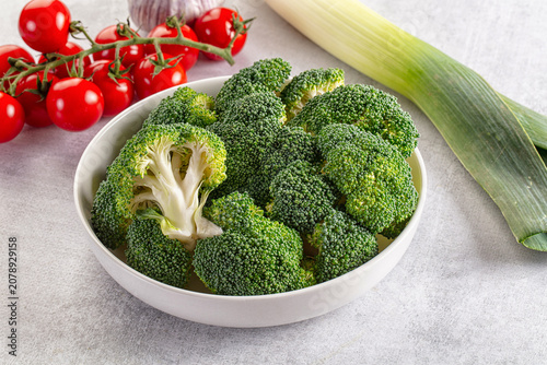 Fresh broccoli florets in white bowl with cherry tomatoes and leek on light gray surface